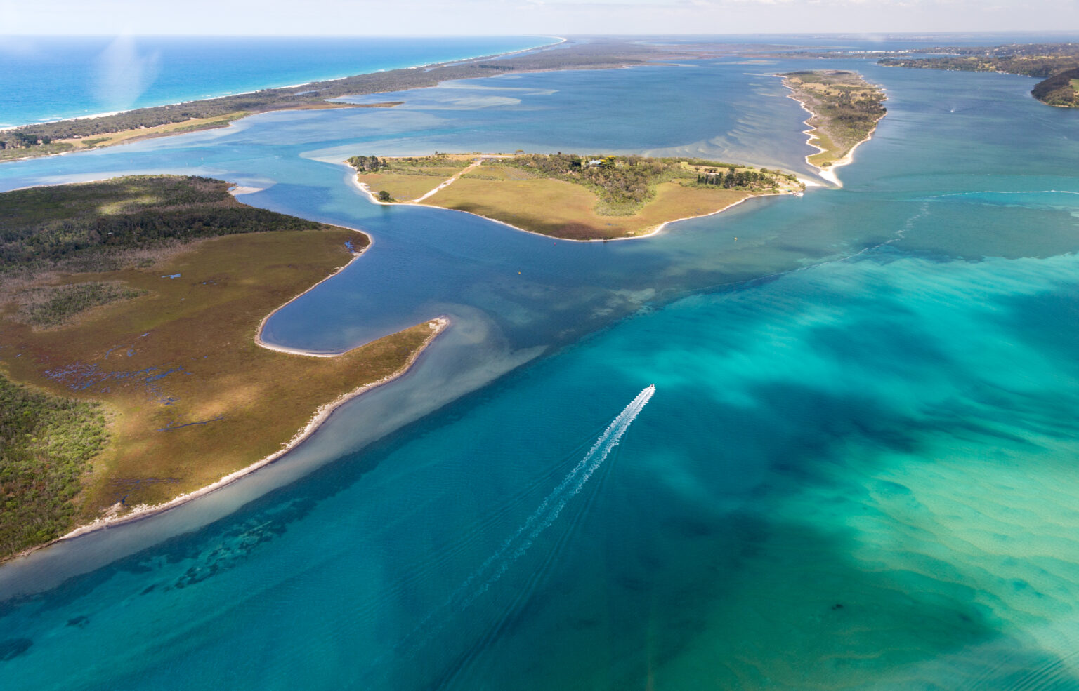 Aerial view of a coastal Gippsland Lakes system with turquoise and deep blue waters, showing a boat leaving a white wake as it moves through the waterway. The scene includes green and brown marshland barrier islands, surrounded by winding channels and bordered by a long, narrow beach - The Ninety Mile Beach - separating the lakes from the open ocean.