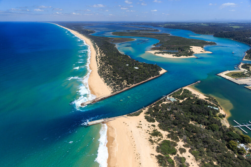 Aerial photograph of an estuary mouth where a Gippsland Lakes meets the ocean at Lakes Entrance, featuring a sandy beach and a long sandspit - The Ninety Mile Beach. The image shows jetties extending into the sea, lush green vegetation along the shoreline, and multiple channels of blue water weaving through low-lying islands and sandbanks.