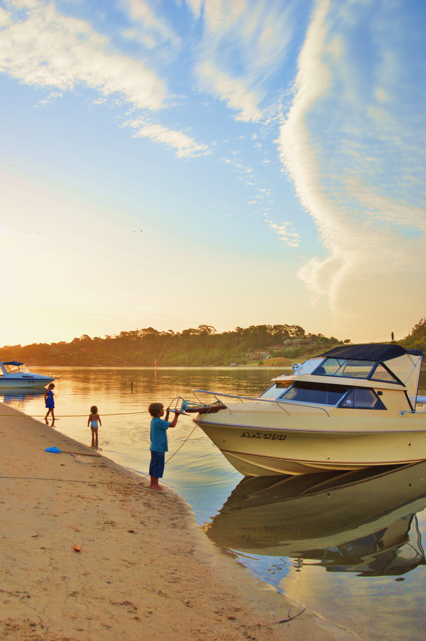 Three children play at the edge of a calm lake at sunset. One child stands on the sandy shore holding a rope attached to a small motorboat, while the other two wade in the shallow water. The sky is filled with wispy clouds, and boats are moored along the lakeside shore.