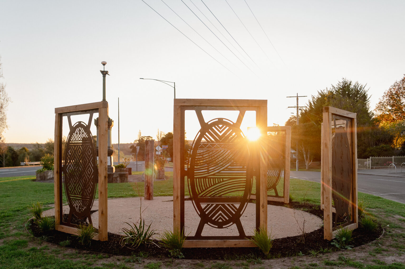 A circular outdoor art installation made up of large wooden frames with intricate metal designs stands in a small park. The sun sets behind the central panel, casting a warm glow and long shadows across the grass and surrounding plants. A road and power lines are visible in the background.
