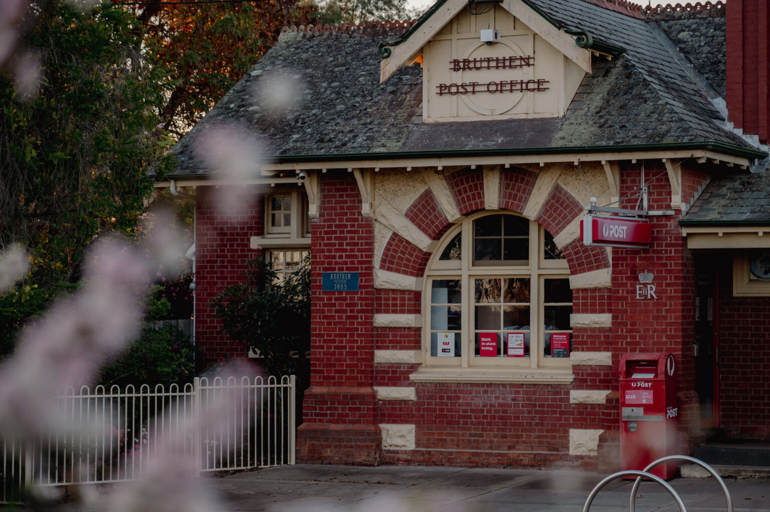 A historic red-brick post office building with cream trim and a slate roof sits behind a white picket fence. The sign above the door reads "Bruthen Post Office." A red Australia Post mailbox and signage are positioned near the entrance, and flowering branches are blurred in the foreground.
