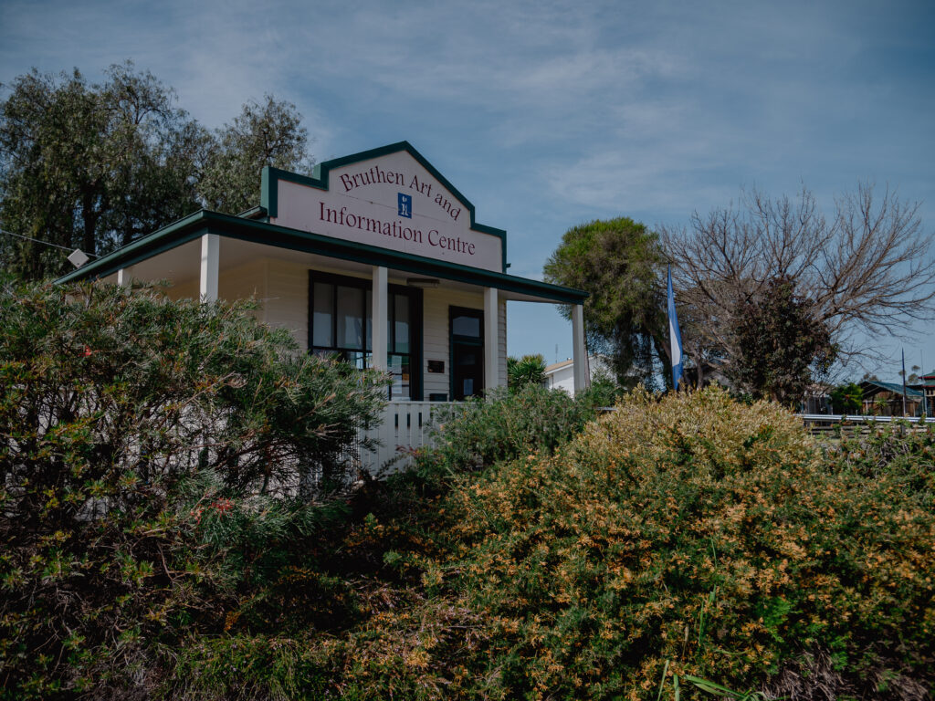 The exterior of the Bruthen Art and Information Centre, a white wooden building with a covered porch and green trim, surrounded by bushes and trees under a blue sky. A sign above the entrance reads "Bruthen Art and Information Centre."