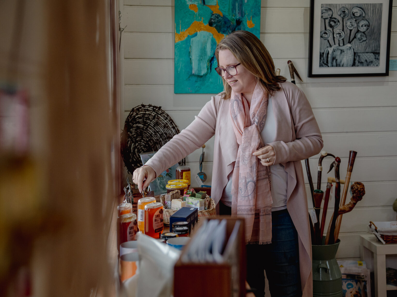 A person stands inside a cozy art and gift shop, reaching toward a table filled with assorted local products such as jams, honey, and crafts. The walls are decorated with colorful paintings and framed artwork, and walking sticks are displayed in a large vase nearby.