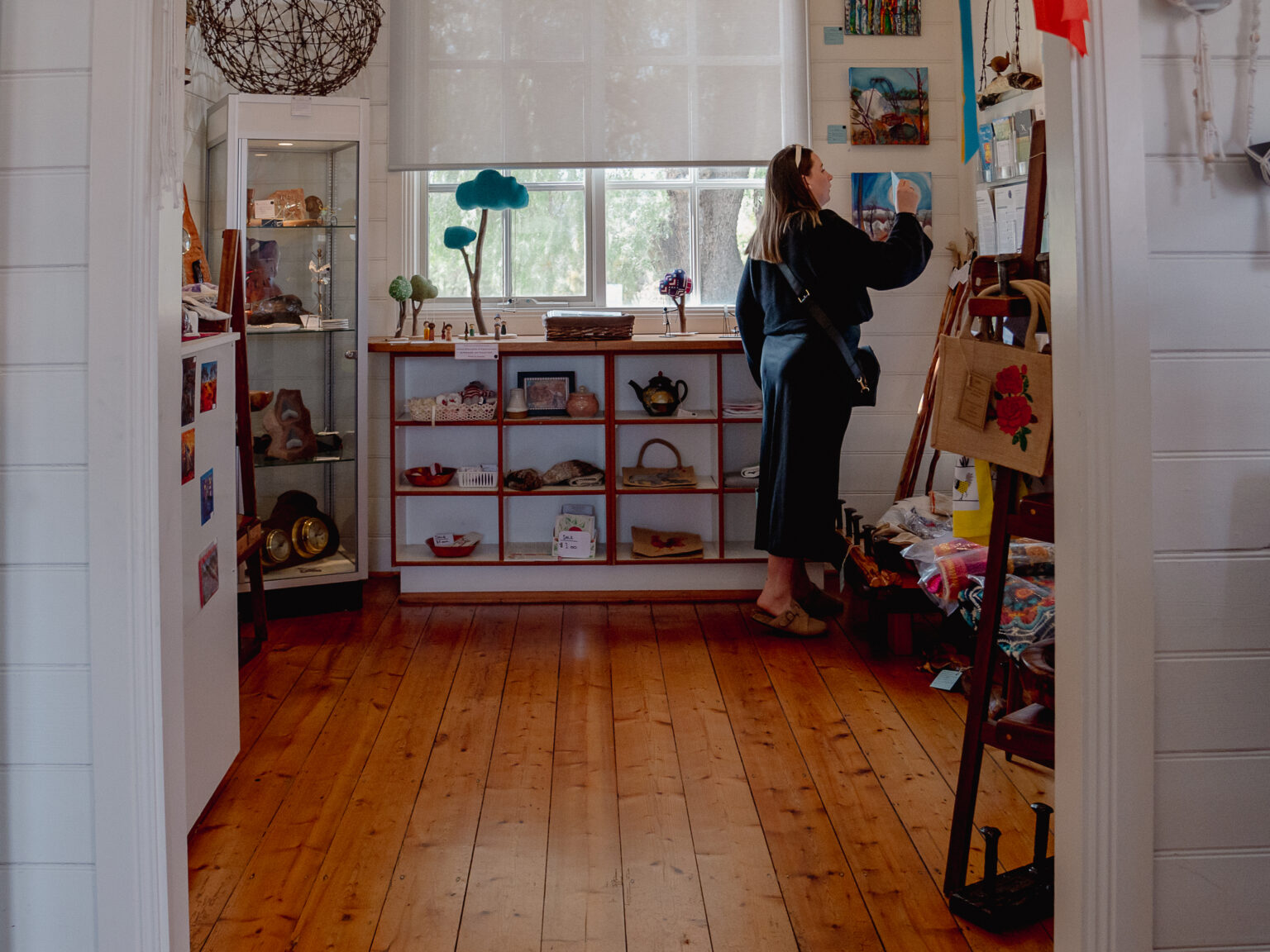 A person browses artwork and handmade items in a small, sunlit gallery space with wooden floors and white walls. Shelves and display cases showcase pottery, jewelry, and crafts, while paintings and photographs hang on the walls.