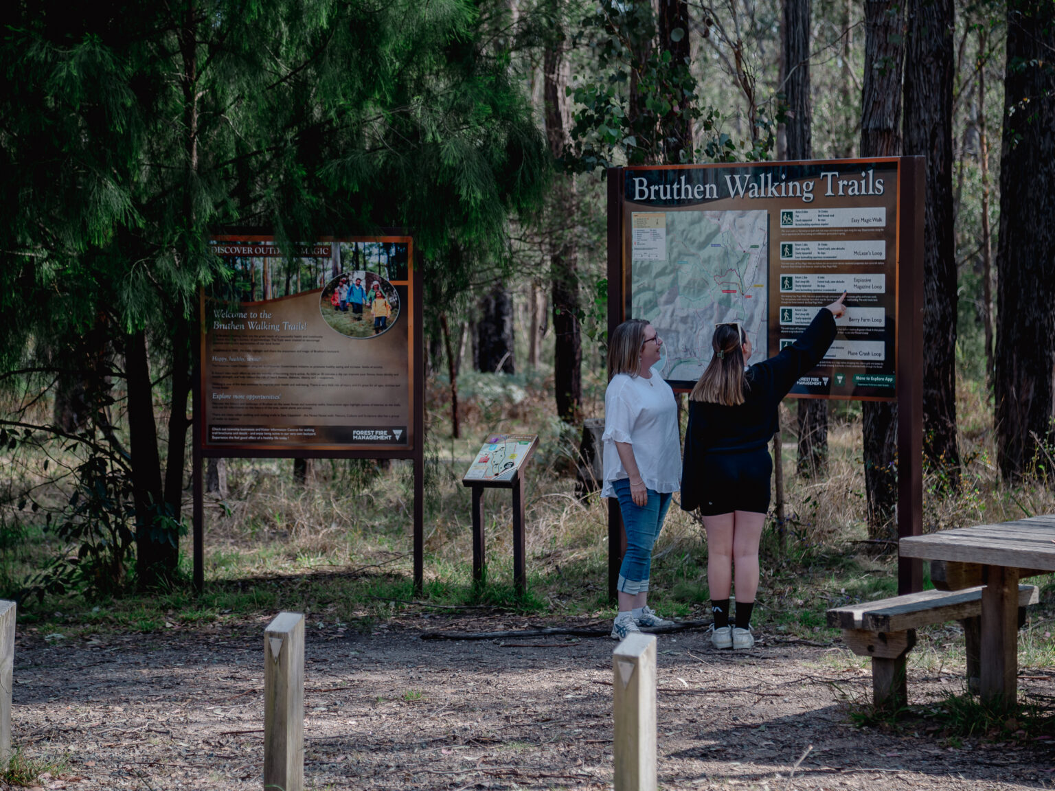 Two people stand in a forest clearing, looking at a large map and signboard for the Bruthen Walking Trails. Tall trees and greenery surround the area, and nearby signs provide information about the walking tracks and local environment.