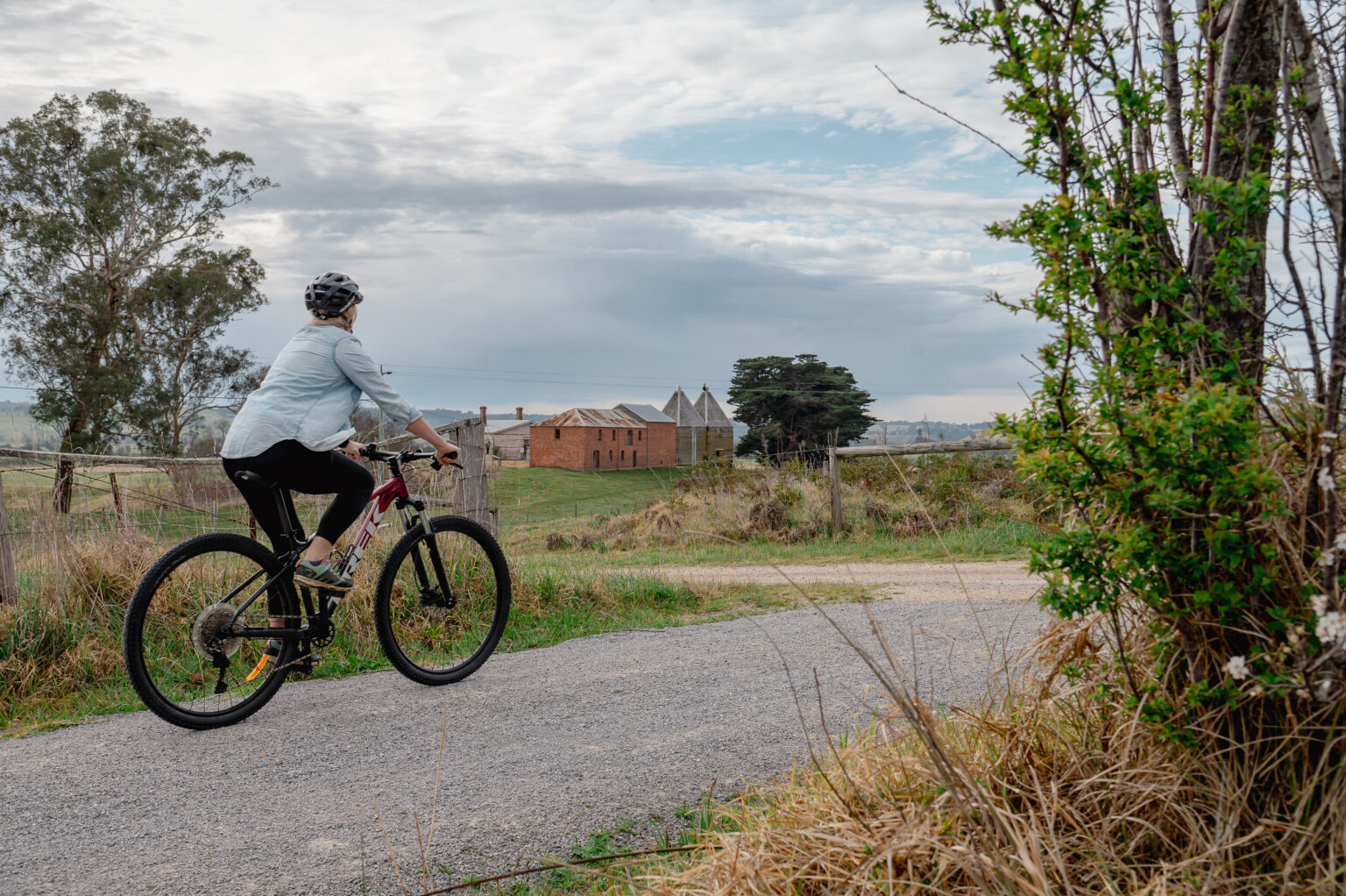 A person rides a bicycle on a paved country path, passing by grassy fields and an old brick building in the distance (Mossiface Hop Kilns). The sky is overcast, and the landscape features scattered trees and a wire fence.