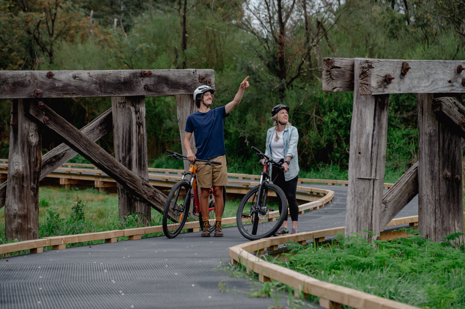 Two people with bicycles pause on a boardwalk path framed by large, rustic wooden beams. One person points ahead while the other looks on, both wearing helmets and casual outdoor clothing.