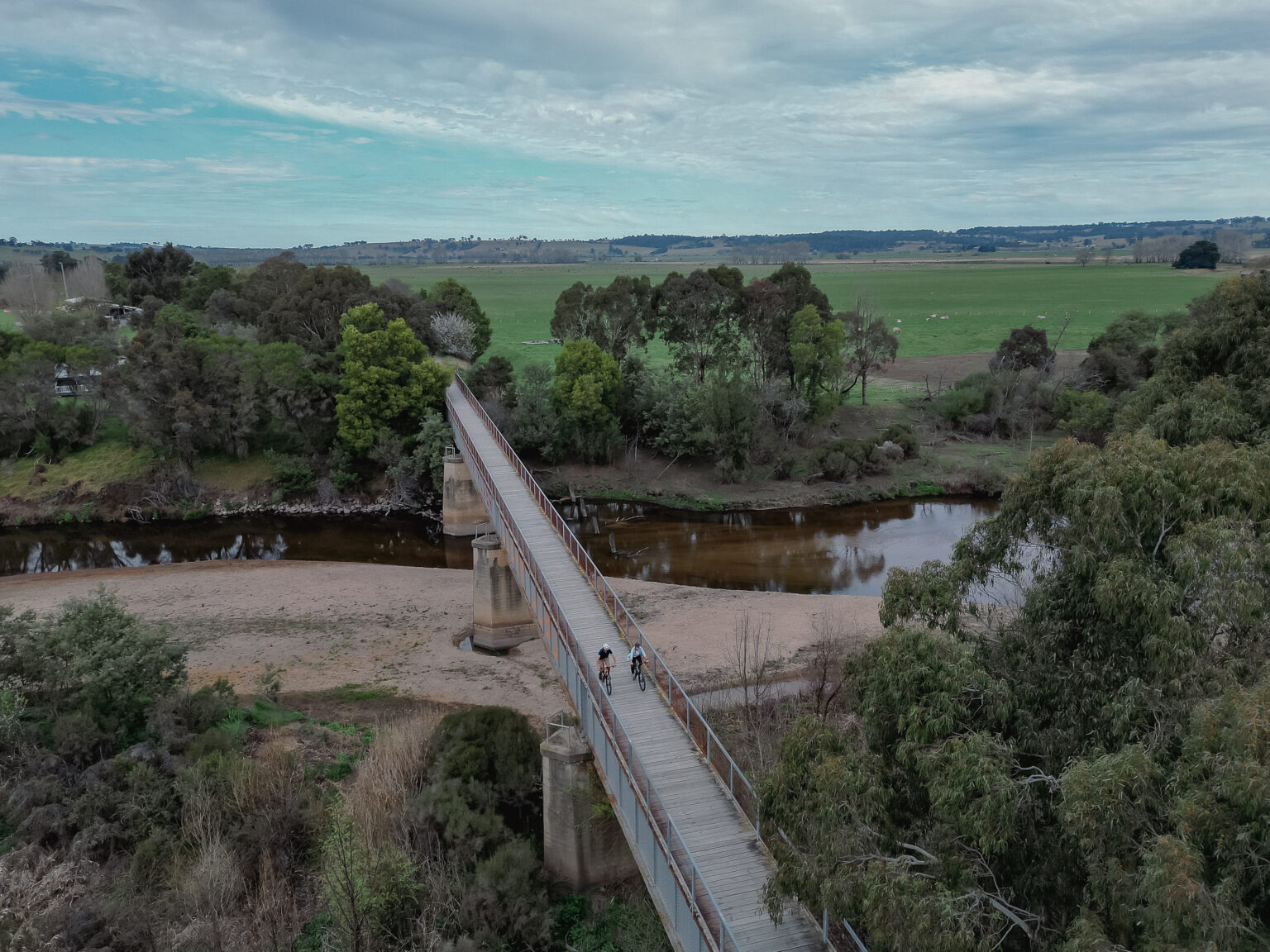 A long, elevated wooden bridge crosses over a river and sandy bank, surrounded by lush green trees and open fields under a cloudy sky. Two cyclists are riding across the bridge.