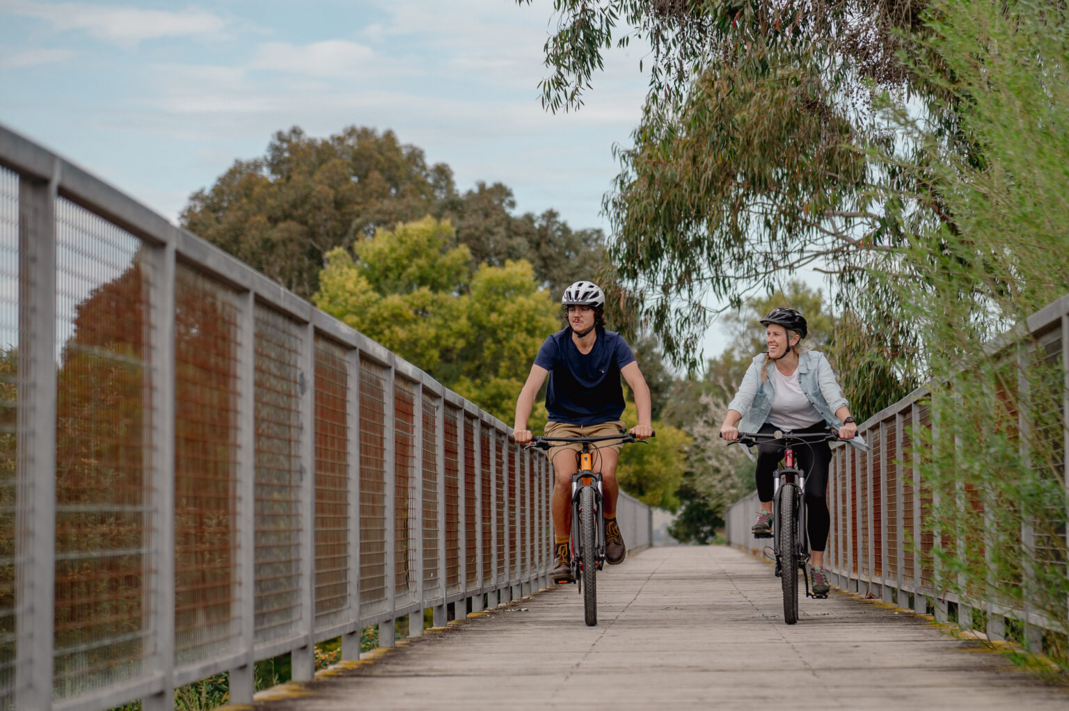 Two cyclists ride across a wooden bridge with metal railings on both sides, surrounded by lush green trees and foliage on a bright day.