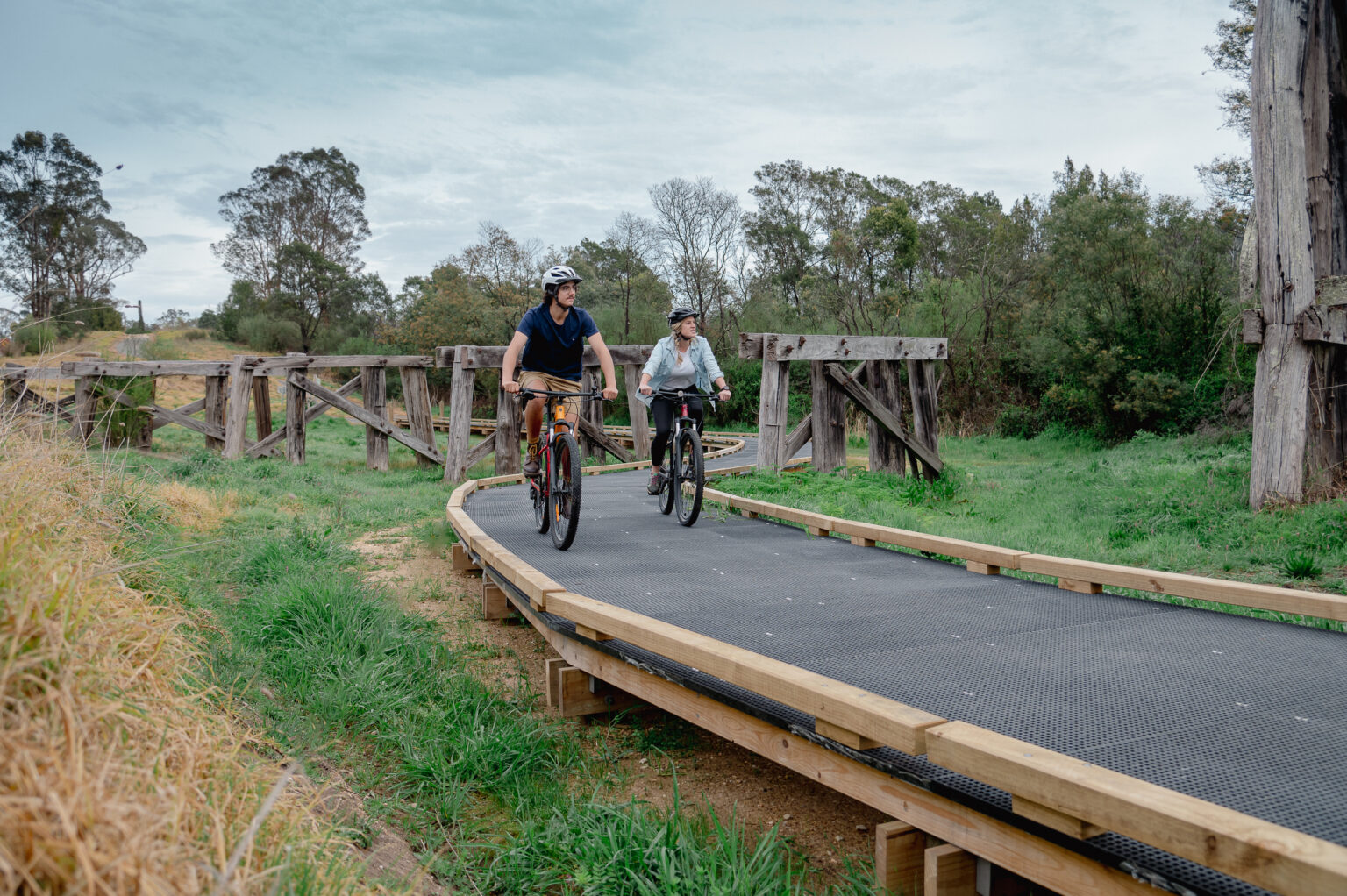 A man and a woman ride their bicycles side by side along a gently curving boardwalk path bordered by grass and old wooden trestle structures, with trees and greenery in the background.