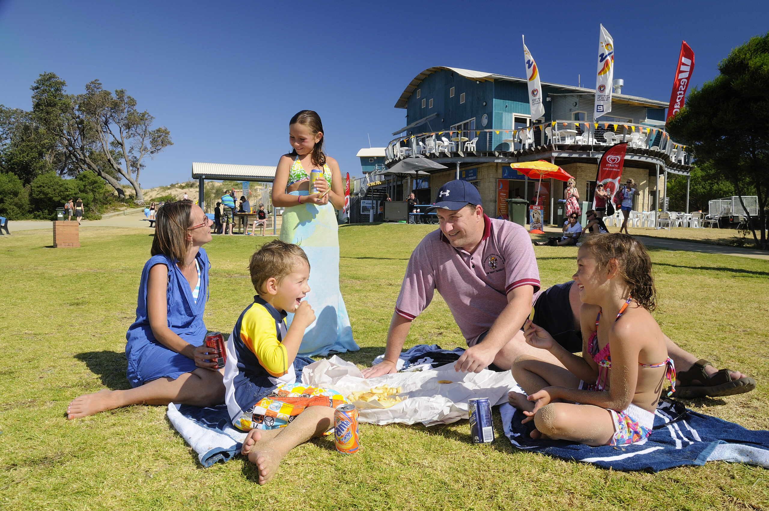 A family, including children and adults, sit together on a blanket spread over green grass, enjoying a picnic with fish and chips. In the background, there is a surf lifesaving club building with outdoor seating, flags, and several people milling about under a clear blue sky.