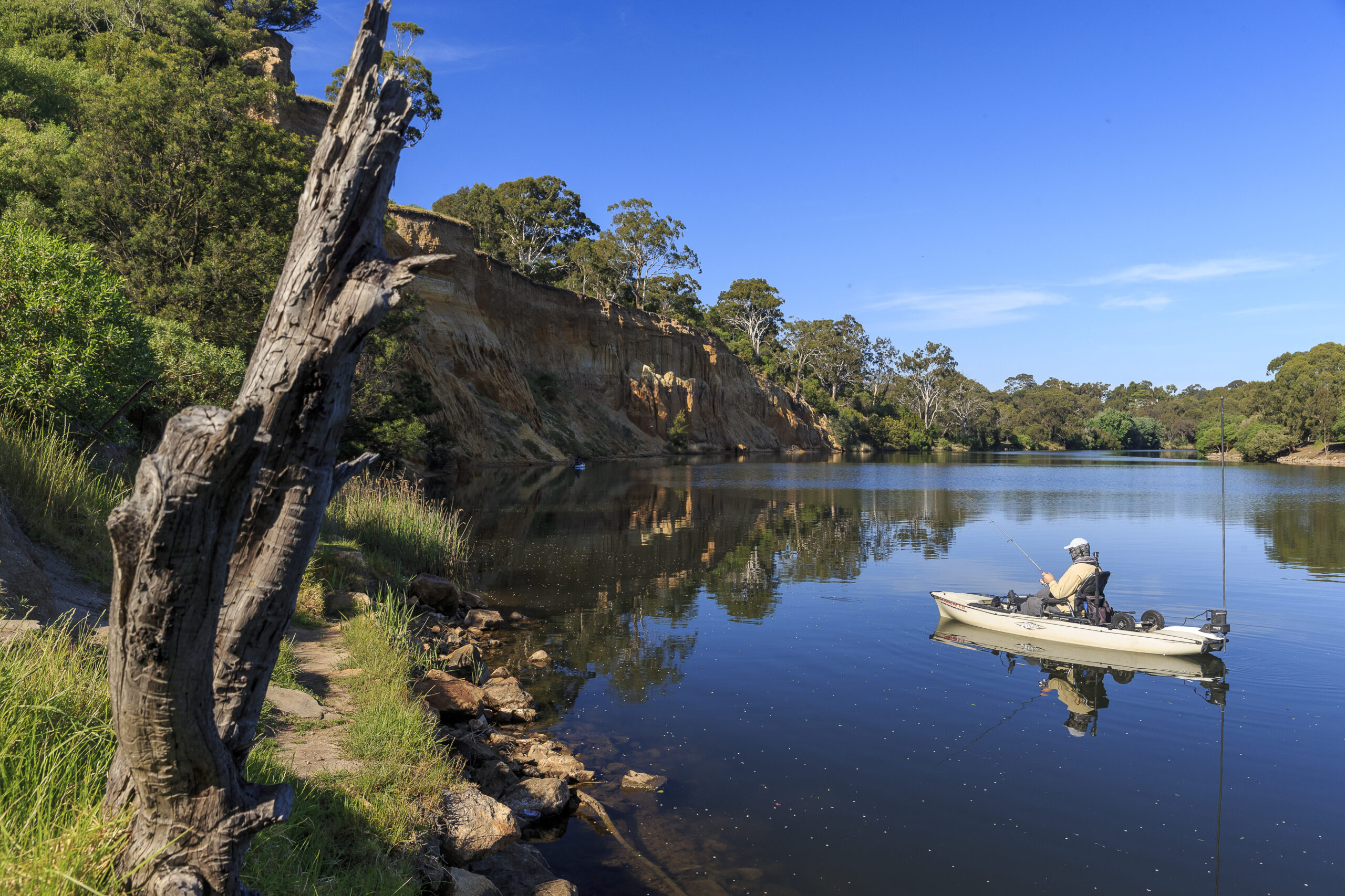 A person sits alone in a small kayak fishing on a tranquil river, surrounded by a steep, tree-lined cliff and lush greenery. The water reflects the blue sky and the surrounding landscape, creating a peaceful, natural setting.