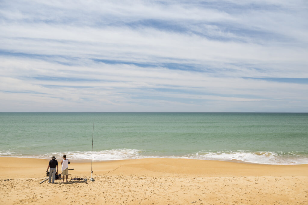 Two people stand on a wide sandy beach next to fishing rods and gear, looking out over calm turquoise ocean waves under a sky streaked with thin clouds.