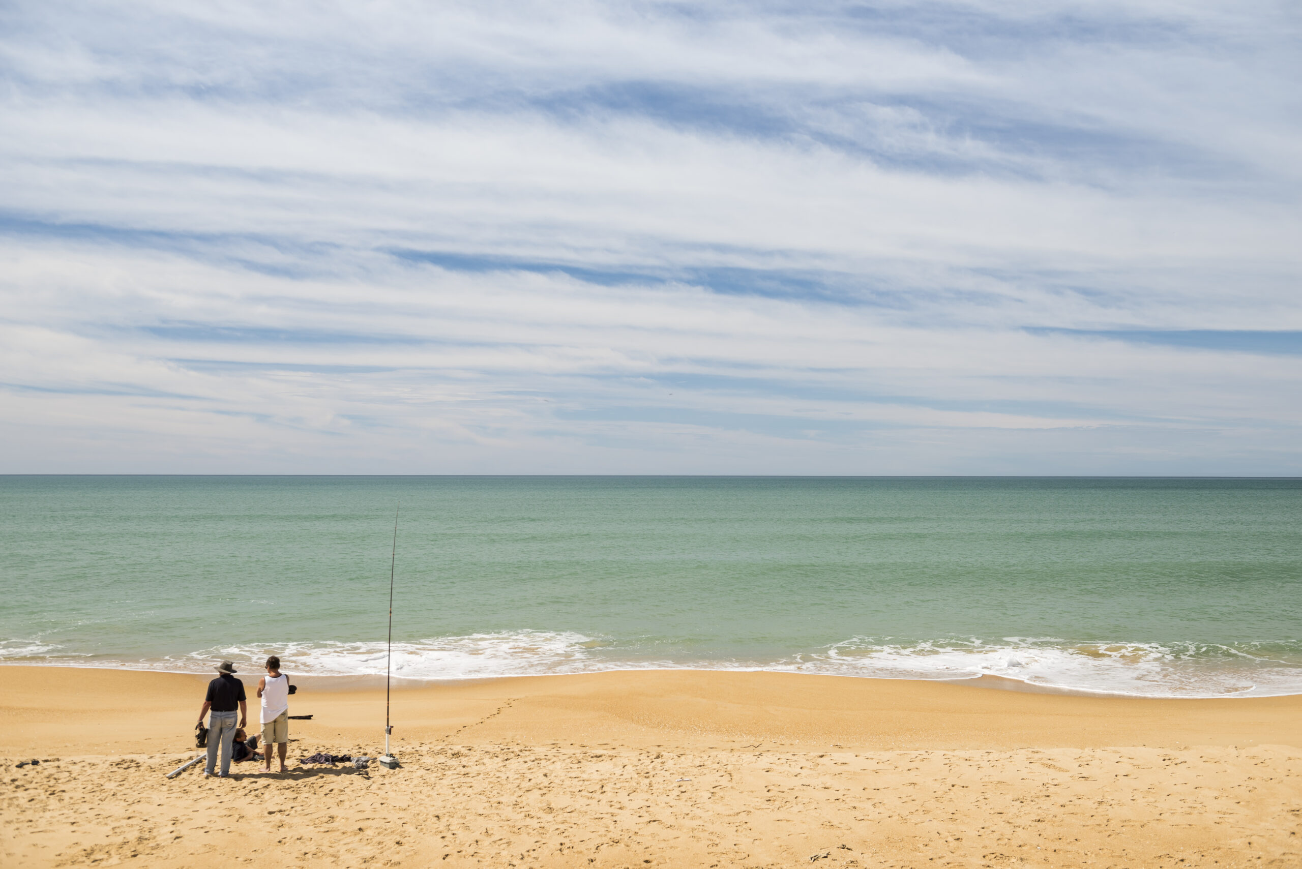 Two people stand on a wide sandy beach next to fishing rods and gear, looking out over calm turquoise ocean waves under a sky streaked with thin clouds.