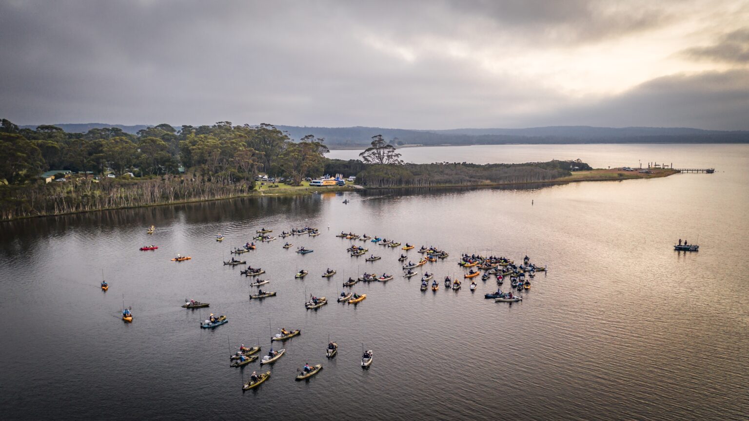 A large group of anglers in colorful kayaks participate in the Hobie kayak fishing tournament on the calm waters of Bemm River, with a forested shoreline and overcast sky in the background.