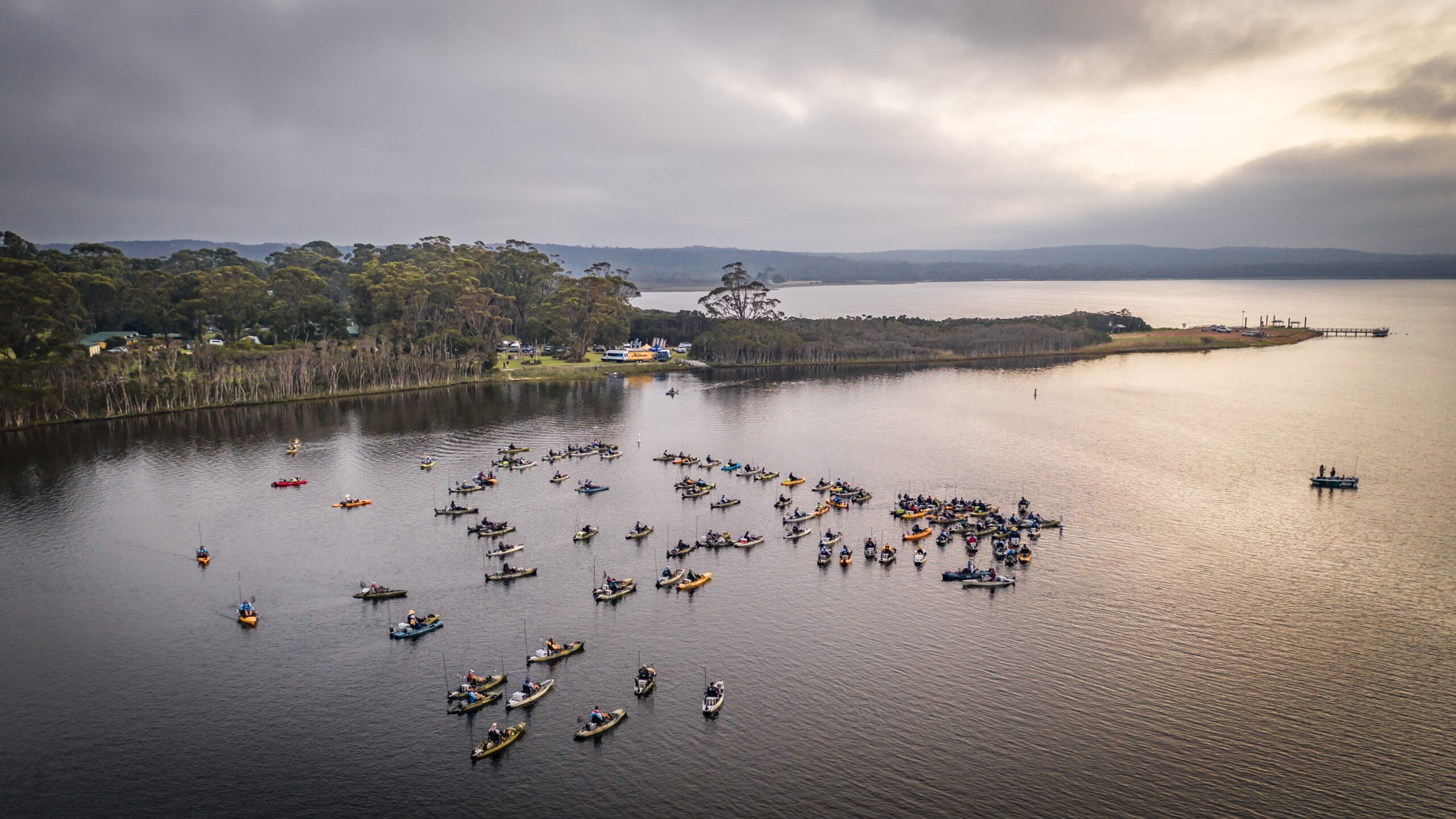 A large group of anglers in colorful kayaks participate in the Hobie kayak fishing tournament on the calm waters of Bemm River, with a forested shoreline and overcast sky in the background.