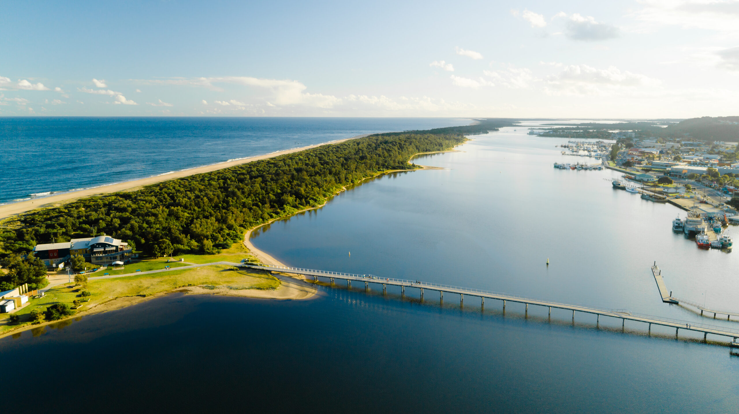 An expansive aerial photograph showing a long, narrow strip of coastal dunes and heath separating a calm inlet from the open ocean. A bridge crosses the water, connecting the shoreline to the town, with boats and buildings visible along the waterfront.