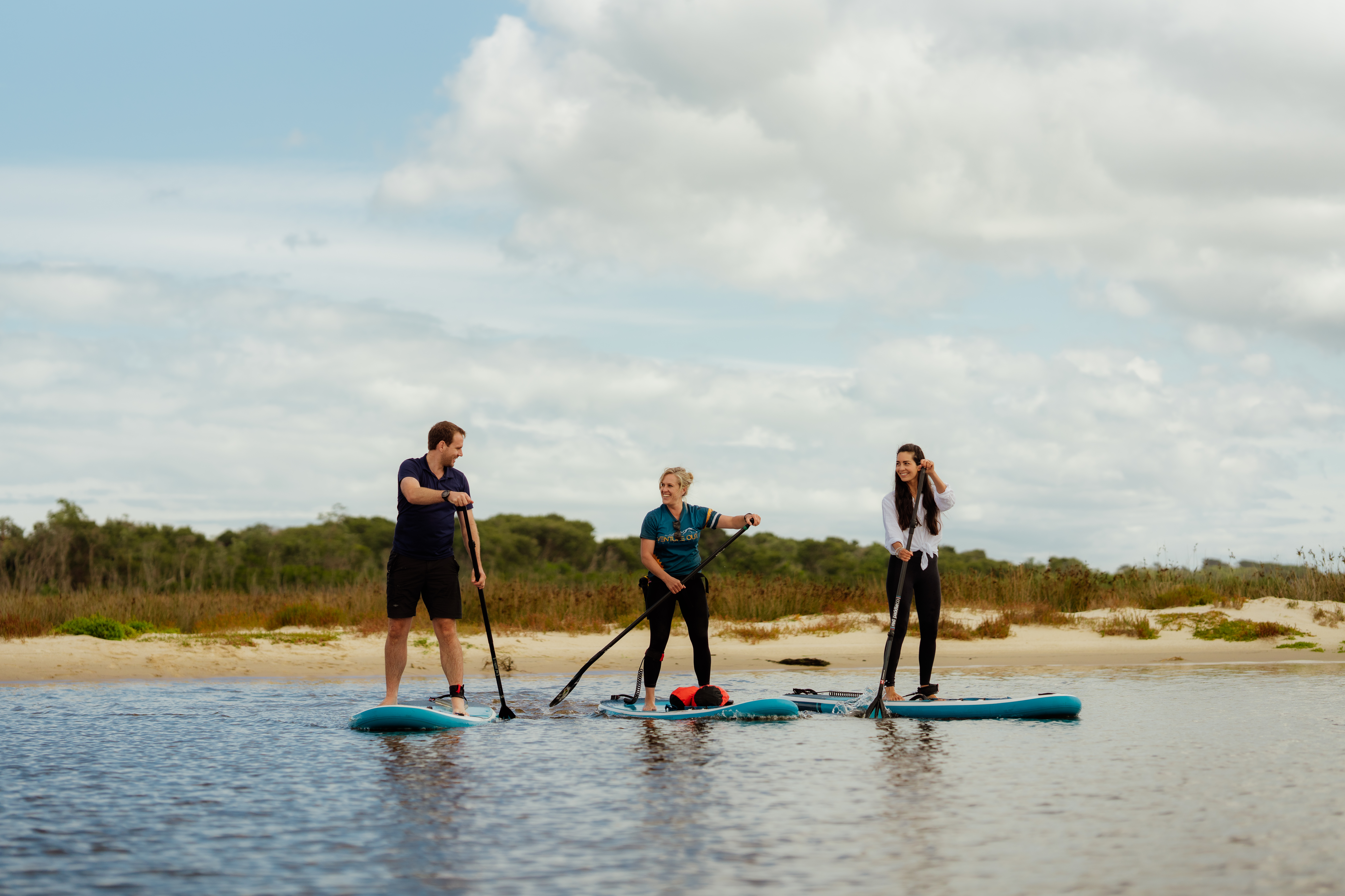 Three people, two women and one man, stand on paddleboards in shallow water near a sandy shore, each holding a paddle. They are engaged in conversation and appear relaxed, with grassy dunes and low vegetation in the background under a partly cloudy sky.