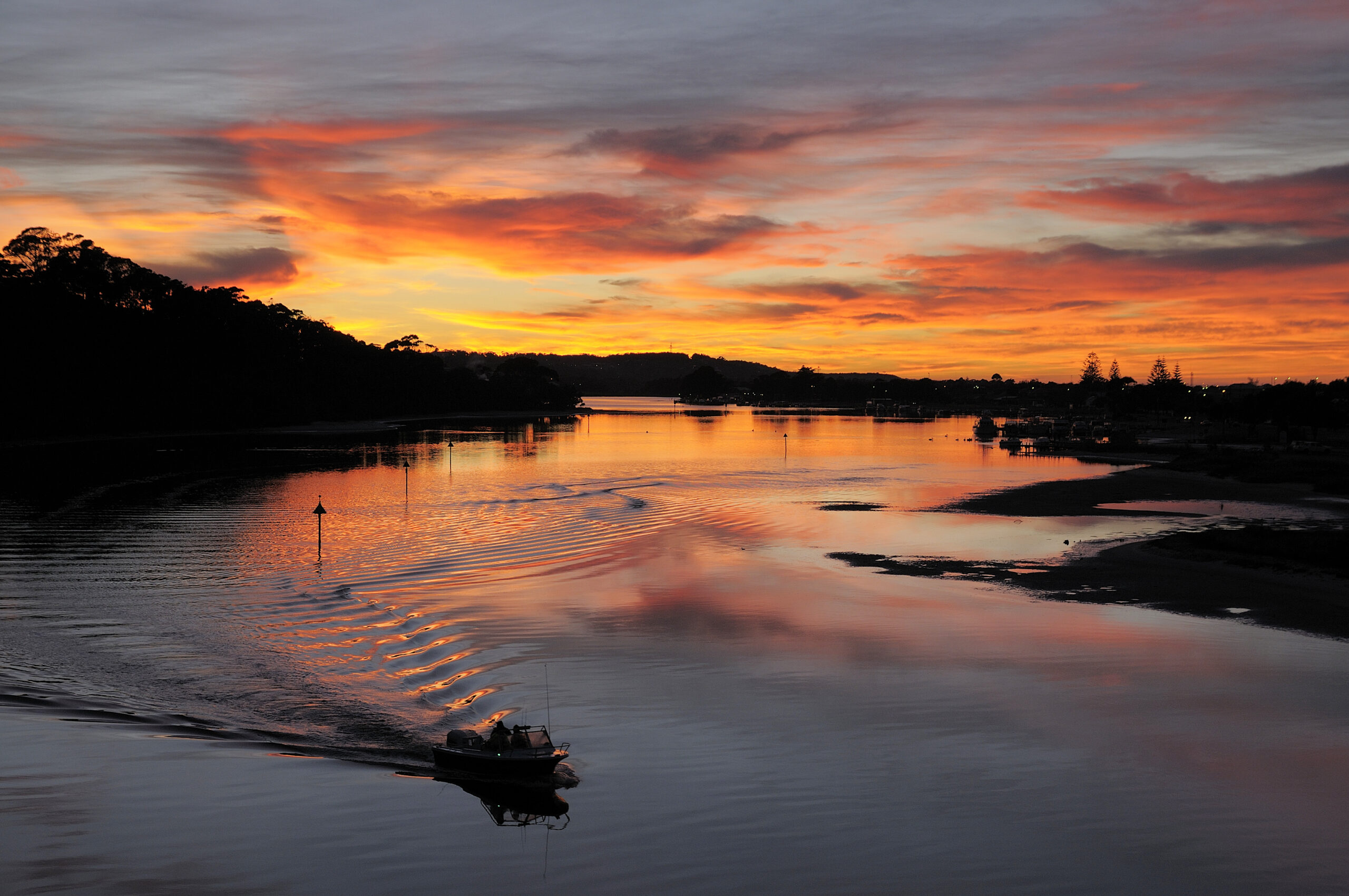 A small motorboat travels along a calm estuary at sunset, leaving rippling waves in its wake. The sky is ablaze with vibrant orange, yellow, and pink hues, which are reflected on the water’s surface. Silhouetted trees and distant hills frame the horizon, while the tranquil scene is mirrored in the glassy river.
