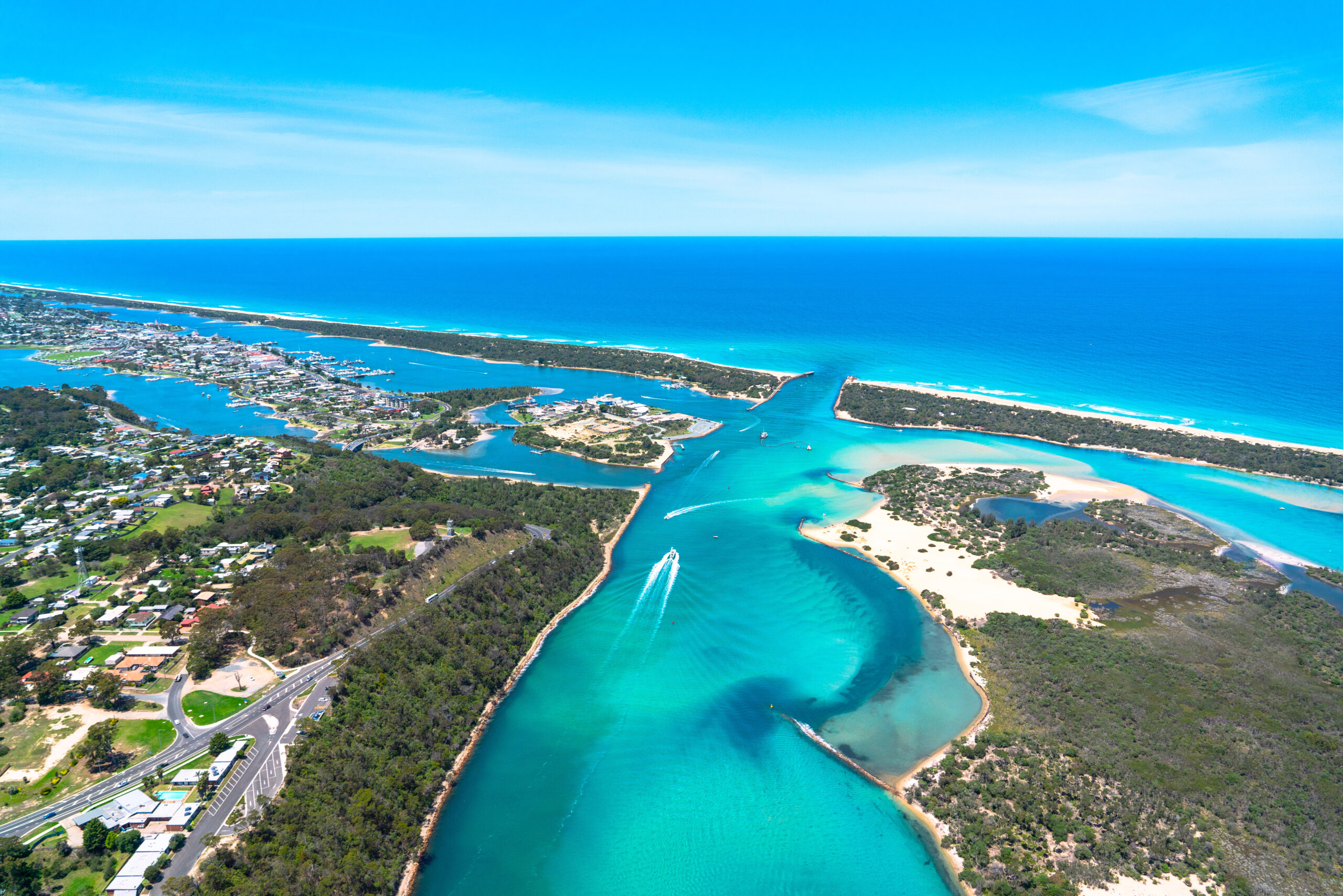 A wide aerial shot of the Gippsland Lakes opening into the ocean, with boats leaving wakes in the clear blue water, sandy beaches, and residential areas visible on the left.