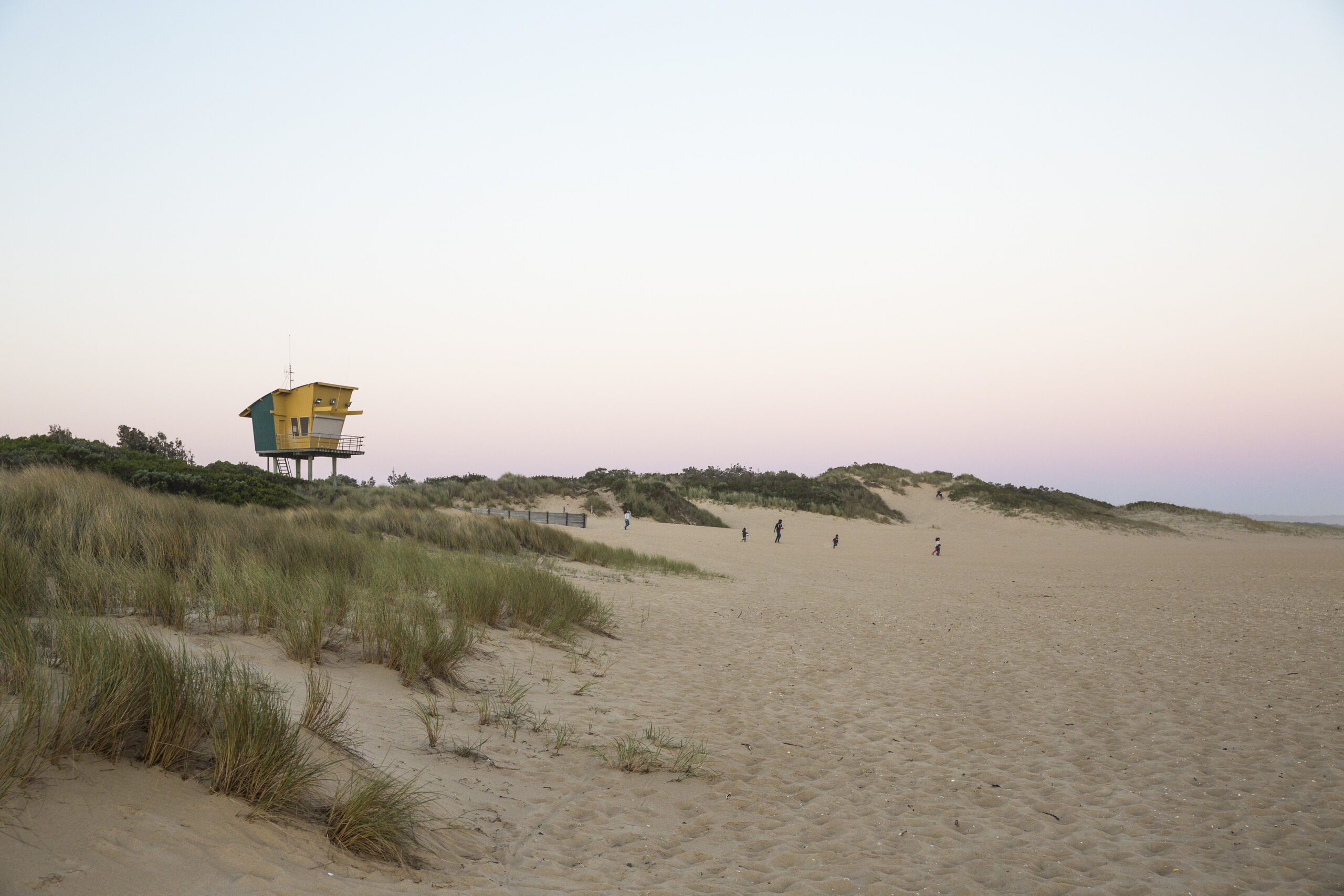 A yellow and blue lifeguard tower stands elevated on stilts among grassy sand dunes. The sky is tinged with soft pastel colors of sunset, and a few small figures walk across the broad sandy expanse in the distance.