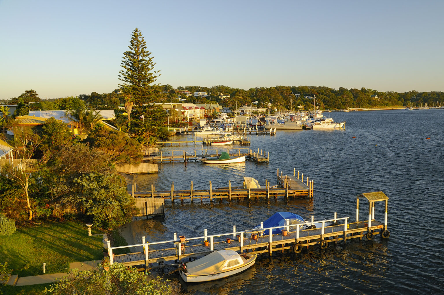 A peaceful waterfront scene at sunset, showing a marina with small boats moored to wooden docks. The shoreline is lined with lush green trees, palm trees, and houses, while the calm water reflects the golden light. In the background, more boats and buildings are visible along the opposite shore.