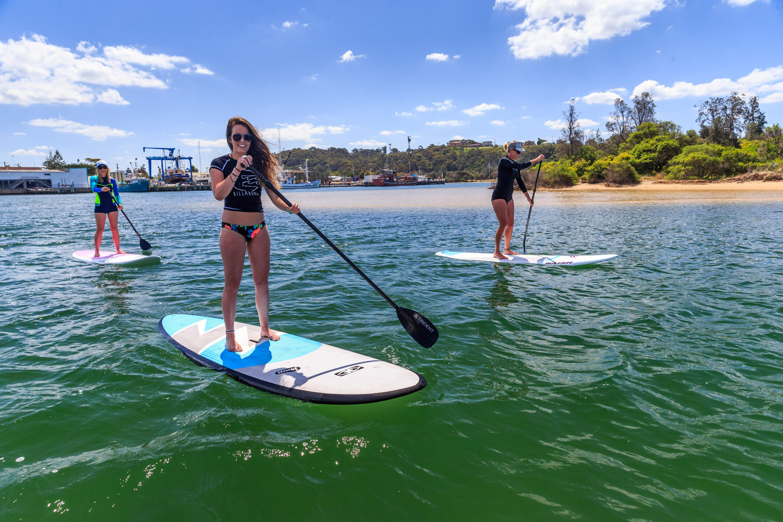 Three women paddleboard on calm blue water in the Cunninghame Arm estuary, with the Lakes Entrance fishing fleet and shore in the background on a sunny day.