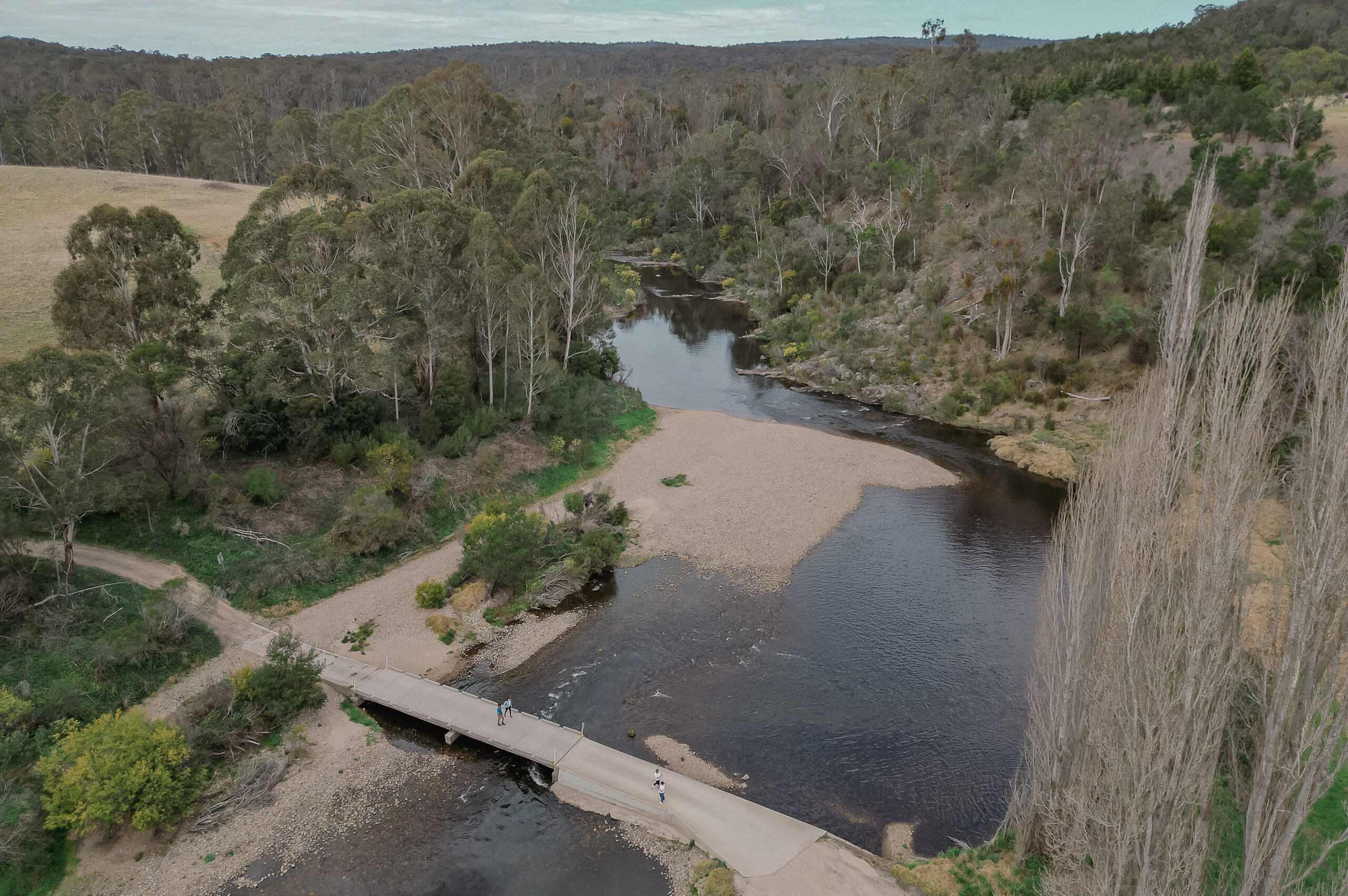 A wide aerial view of the Tambo River winding through a forested landscape. A small bridge crosses the river, with a few people visible on it. The scene includes grassy banks, pebbled shores, and dense trees extending into the background.