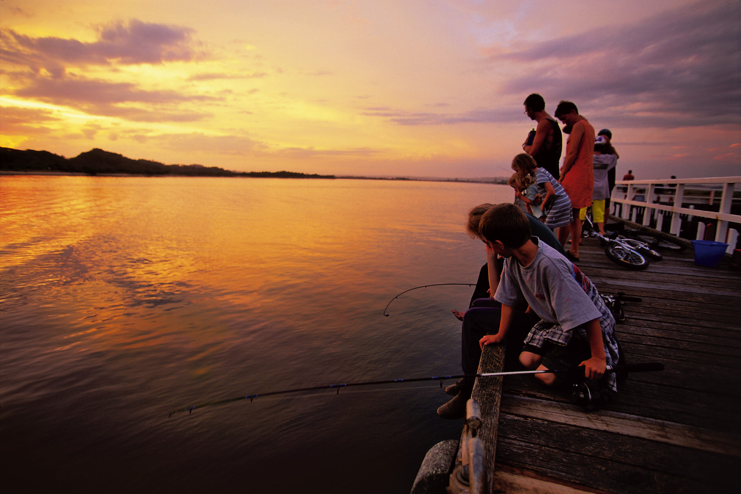A group of children and adults gather on a wooden jetty at sunset, fishing over the calm water. The sky is filled with warm hues of orange and pink, and the water reflects the colors of the sunset. Bicycles and a blue bucket are placed on the pier, and the distant shoreline is visible under the glowing sky.