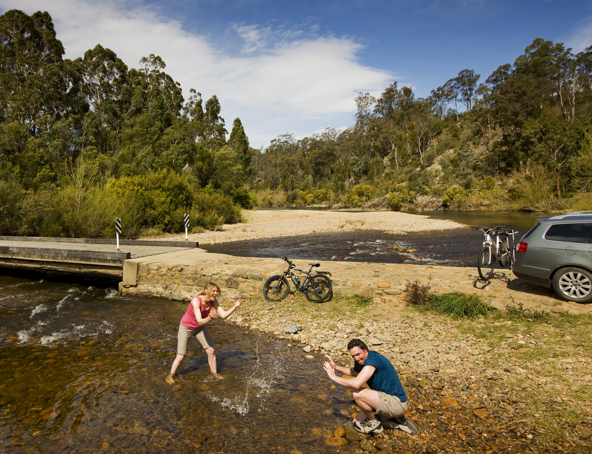 A woman stands in a shallow, rocky river stream playfully splashing water toward a man who is crouched on the shore. Two bicycles and a car are parked nearby, with a low bridge and dense green forest in the background under a blue sky.