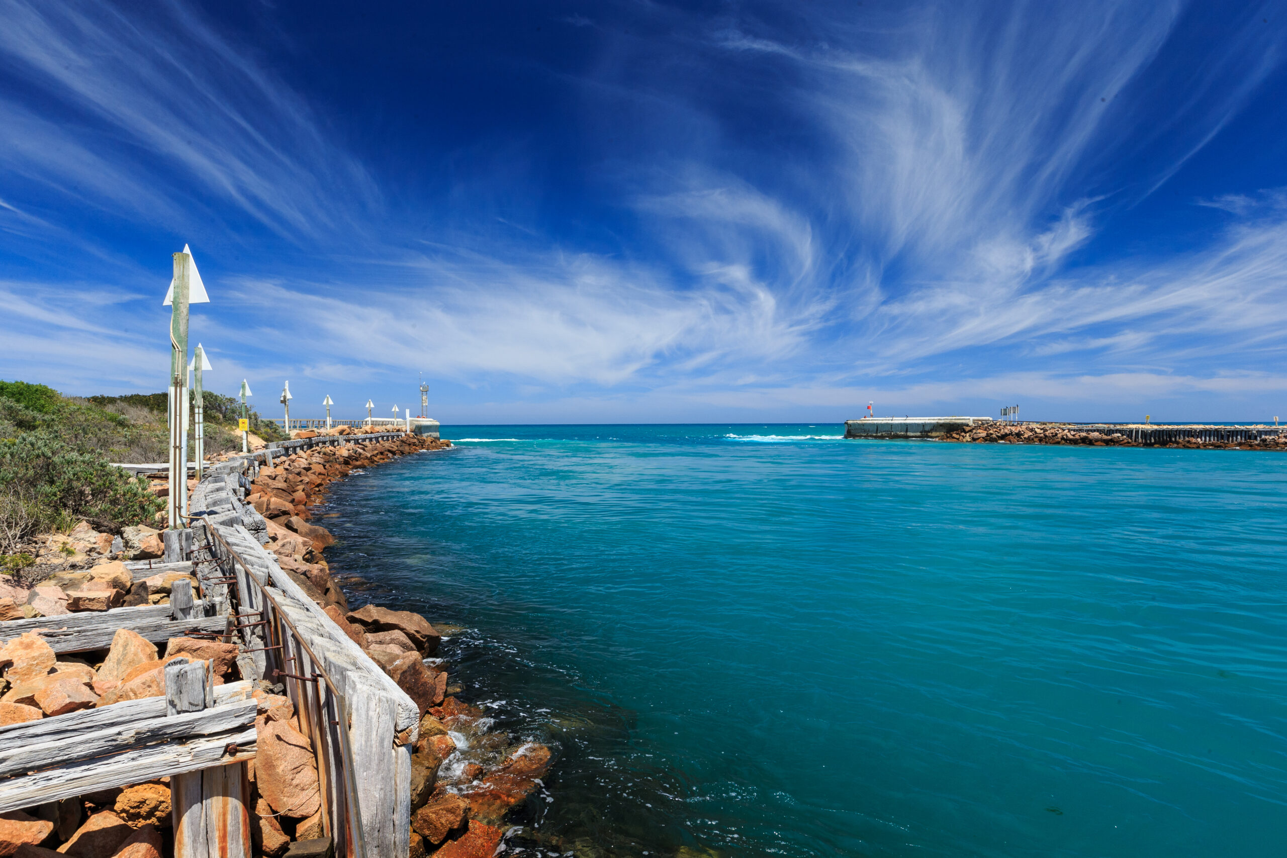 A rocky breakwater extends into a bright blue sea under a vivid blue sky with wispy clouds. Wooden railings line the path along the rocks, and white markers are spaced along the breakwater. The water is clear, and the horizon meets the open ocean beyond the entrance to a sheltered harbor.