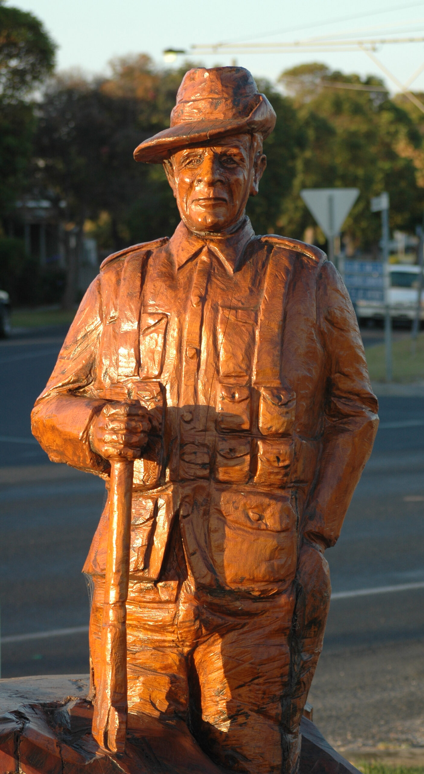 A life-sized wooden sculpture of a soldier in uniform stands outdoors near a street intersection, holding a walking stick and wearing a hat, with trees and road signs visible in the background.