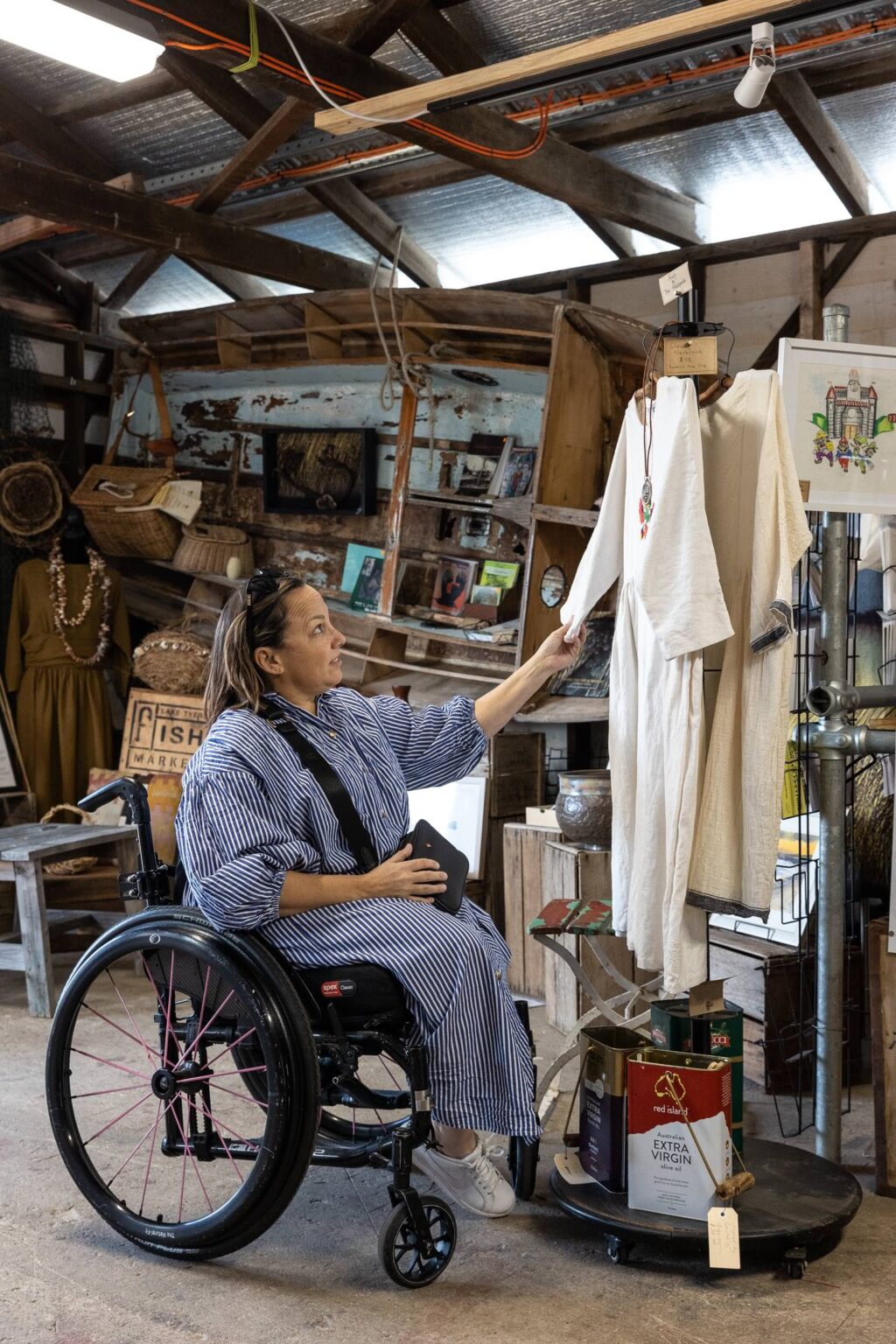 A woman in a wheelchair is inside a rustic shop filled with vintage and handmade items. She holds a crossbody bag and reaches out to touch a cream-colored dress hanging on a rack. The shop features wooden shelves, baskets, and various crafts and decorations on display.