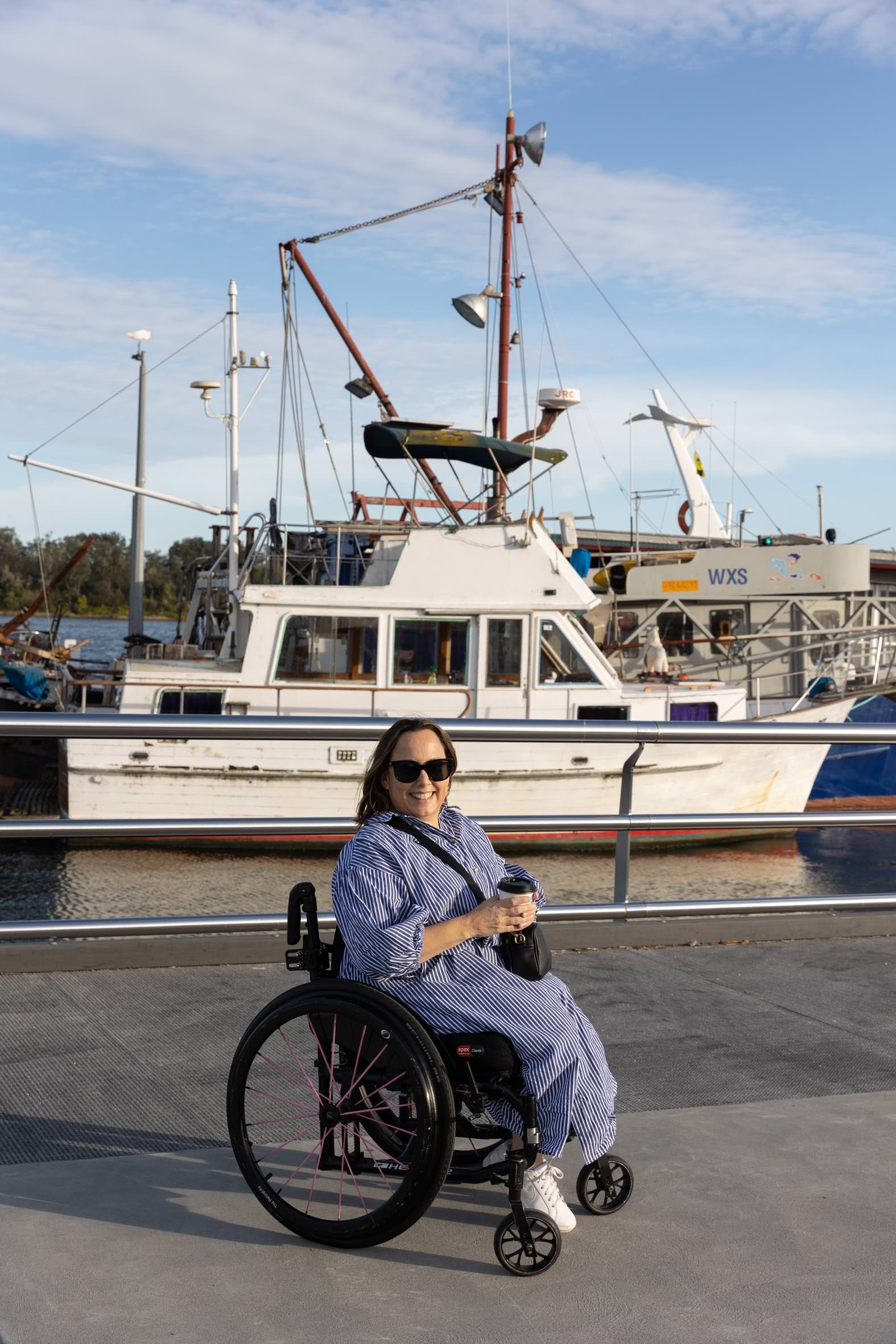 A woman in a wheelchair relaxes on a waterfront promenade, holding a drink. Behind her, several fishing boats and yachts are docked in the marina, with a clear blue sky and calm water creating a scenic backdrop.