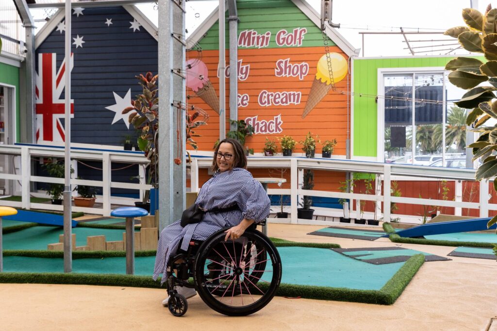 A woman in a wheelchair explores a vibrant mini golf course with smooth, accessible paths. The surroundings include colorful buildings decorated with large painted flags, stars, and playful signage, as well as lush potted plants and mini golf obstacles.