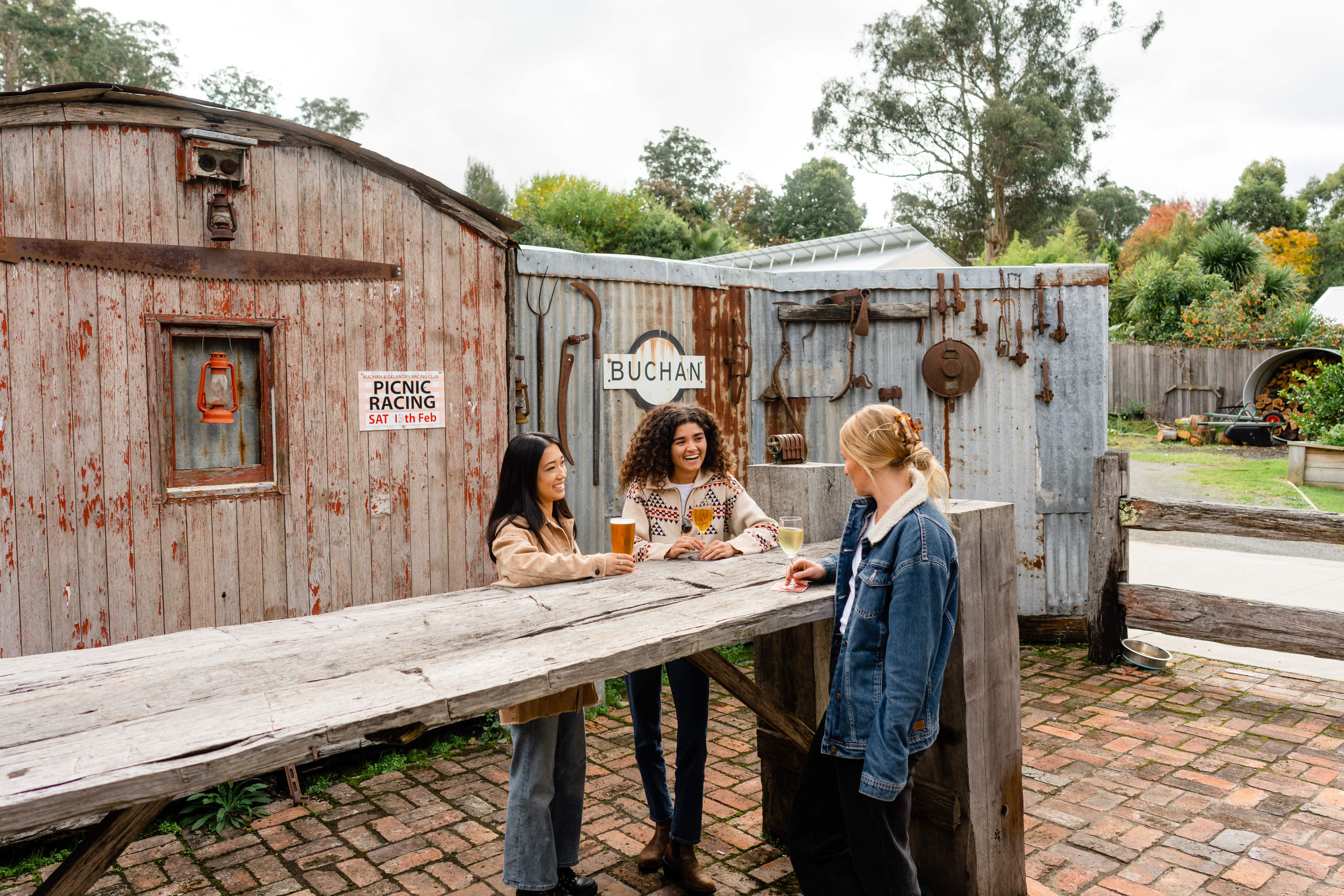 Three women stand and sit around a rustic wooden outdoor table in front of weathered, corrugated iron and timber sheds. The setting includes old tools hanging on the wall, a lantern in a small window, and a sign reading "BUCHAN." The atmosphere at the Buchan Caves Hotel is casual and relaxed, with drinks in hand, surrounded by greenery and a brick-paved ground.