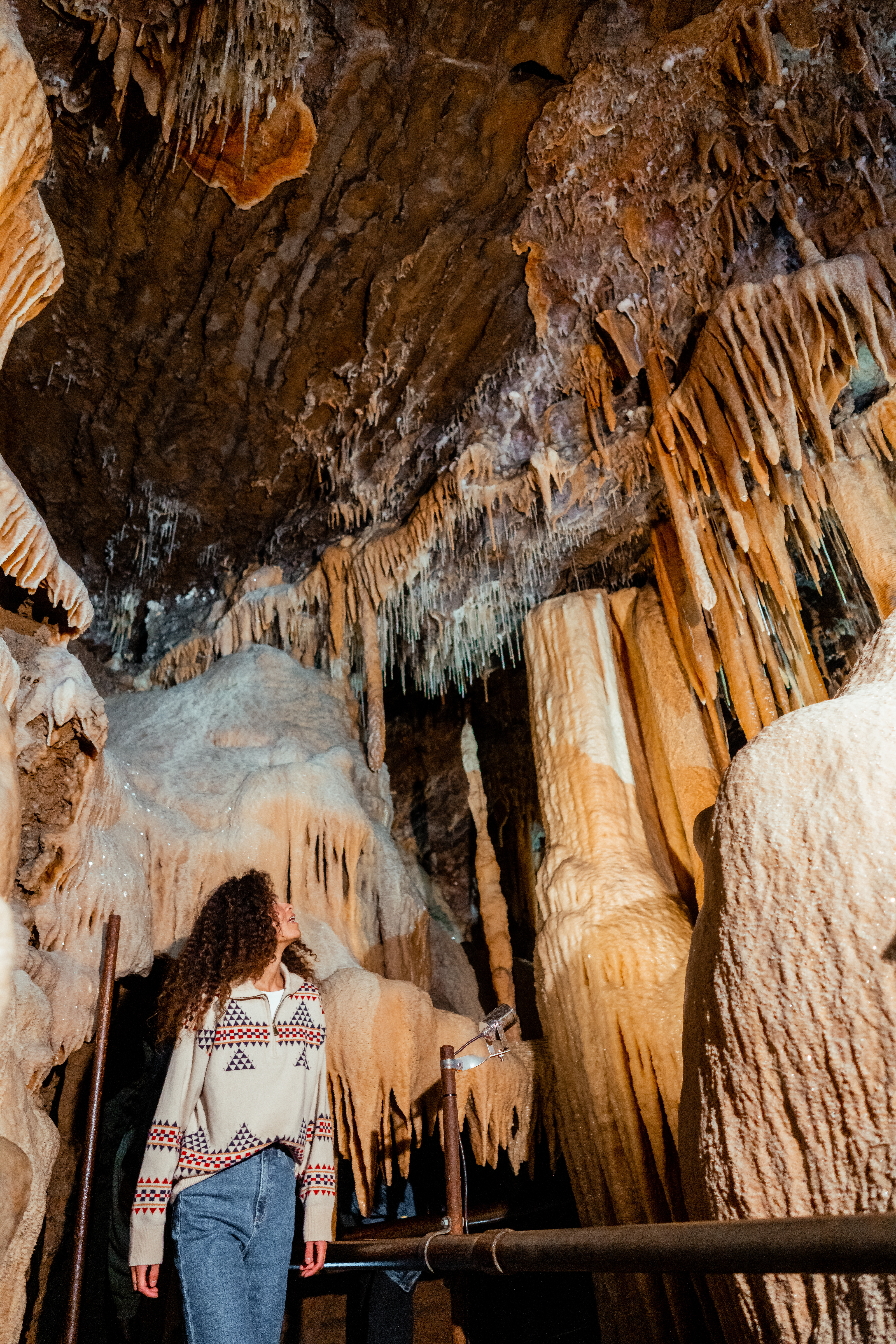 A young woman with curly hair, wearing a patterned sweater and jeans, stands inside a cave surrounded by dramatic formations of stalactites and stalagmites. The individual looks up in awe at the intricate mineral deposits hanging from the cave ceiling, illuminated by soft artificial lighting.