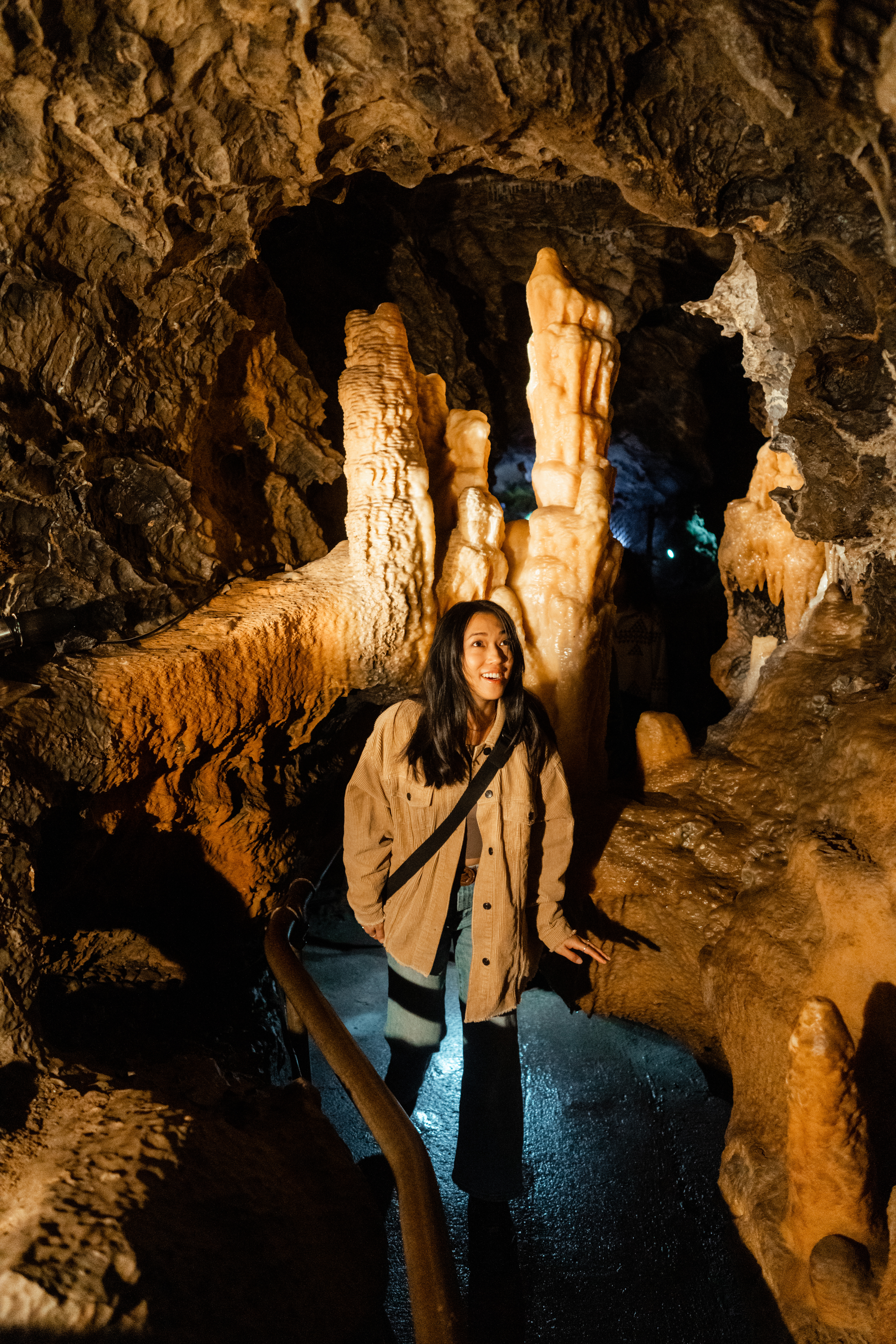 A woman with long dark hair, dressed casually in a tan jacket and jeans, explores the inside of a cave. She smiles as she walks along a path bordered by railings, surrounded by large, orange and white mineral formations and textured cave walls.