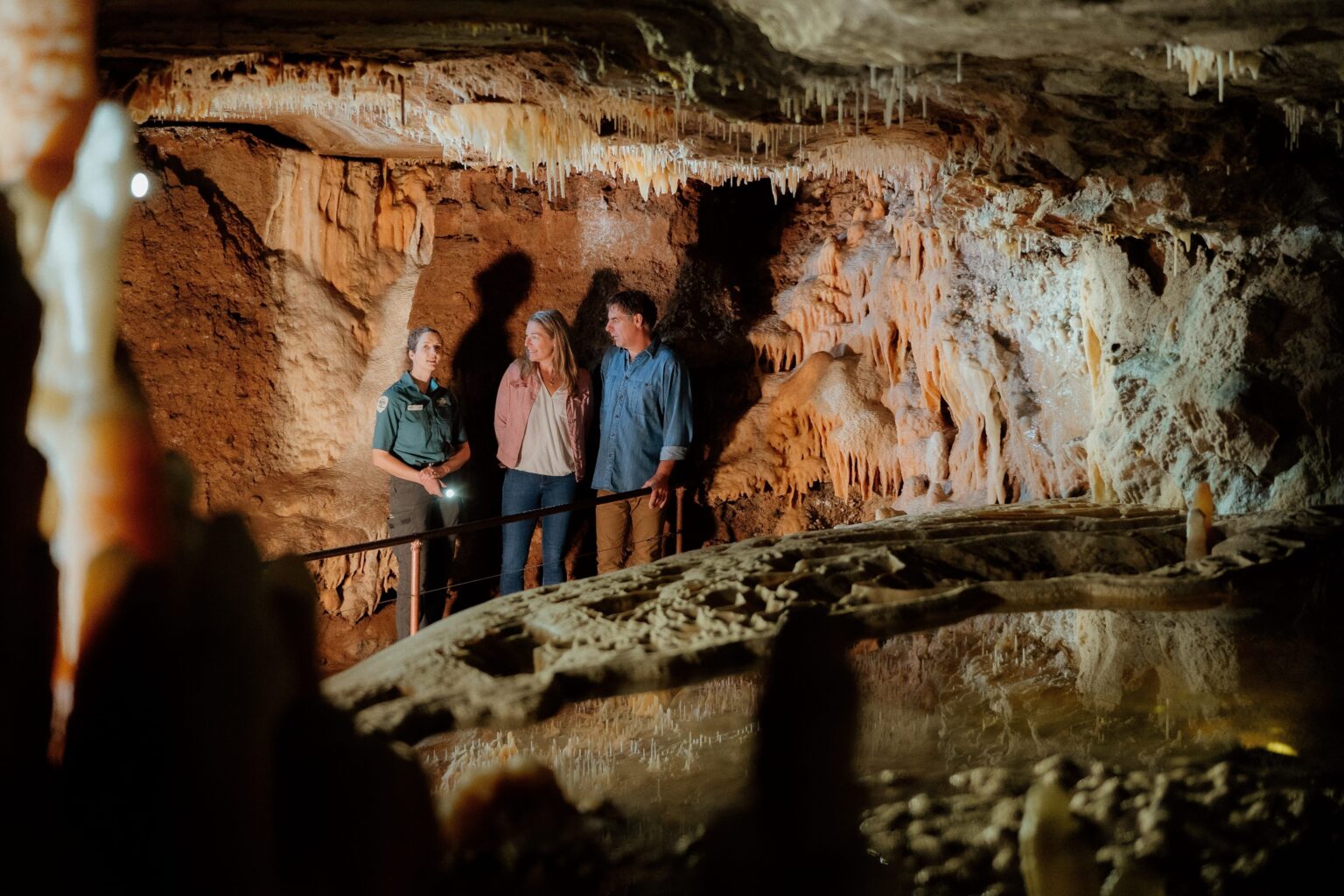 Three people, including a park guide, stand inside a spacious cave chamber. The group listens attentively as the guide explains features of the cave, which is filled with striking stalactites and stalagmites. The scene is warmly lit, highlighting the natural beauty and textures of the cave interior.