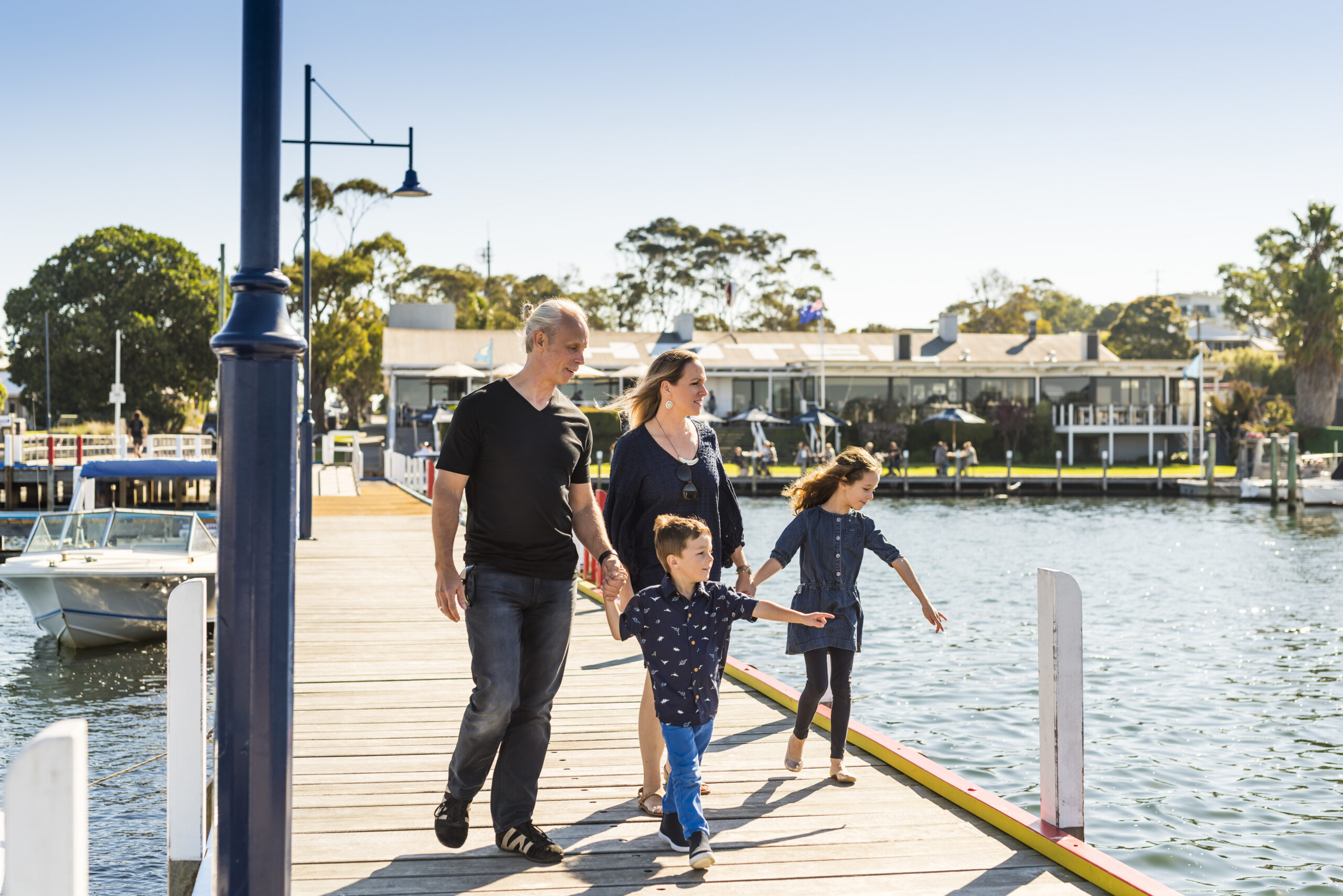 A family of four, including two adults and two children, walks together on a wooden pier by the water on a sunny day. A waterfront pub "Metung Hotel" and picnic tables can be seen in the background across the water.