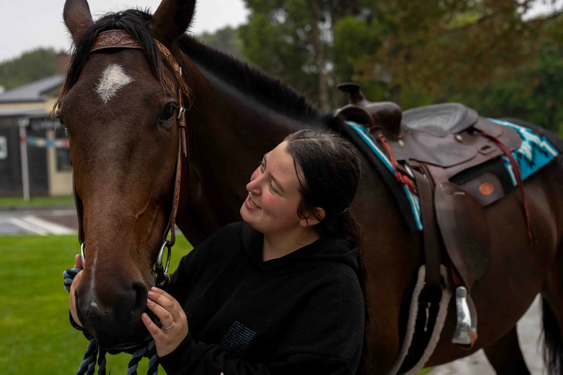 Smiling woman standing close to a saddled bay horse with a white blaze